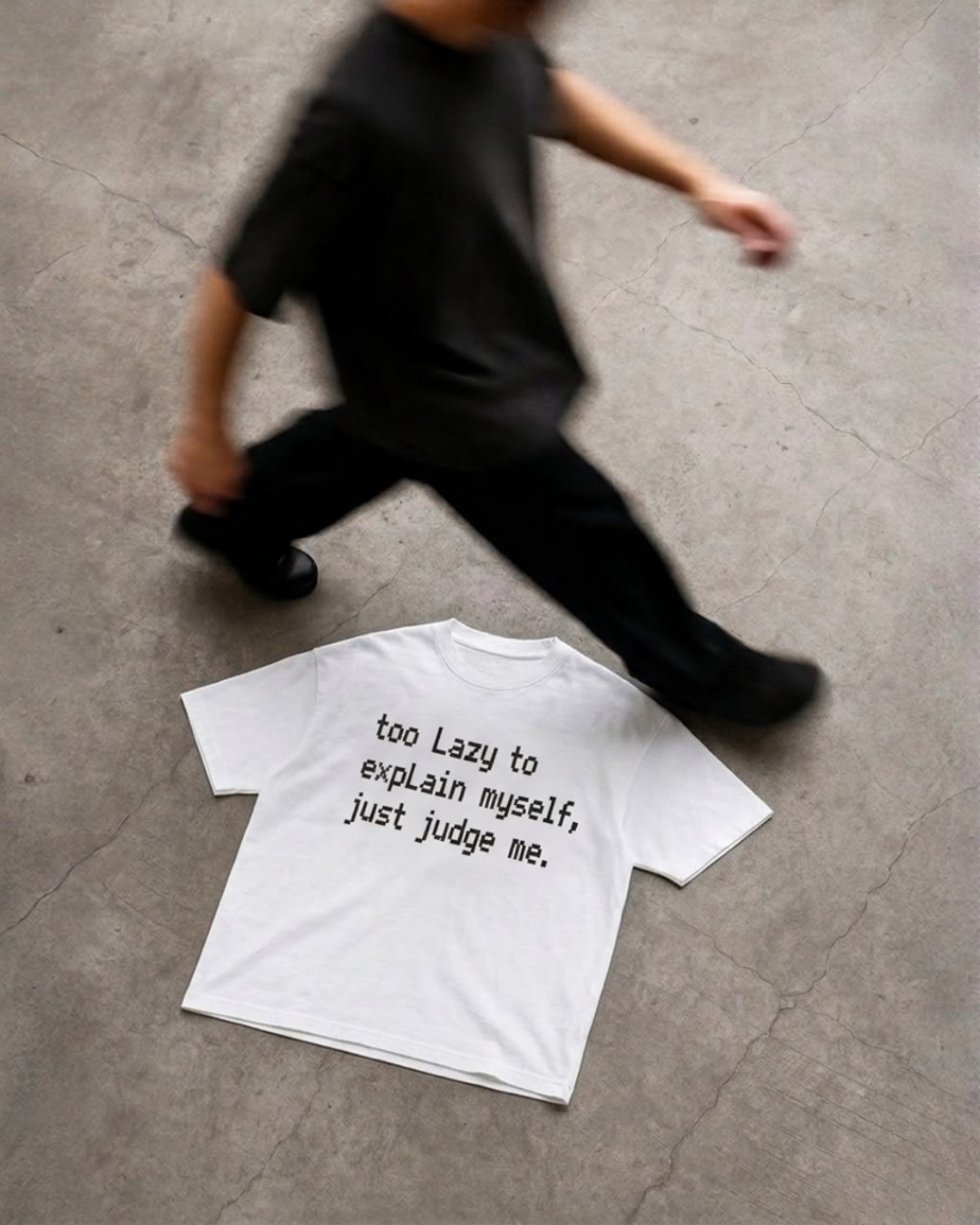 A white short-sleeve T-shirt with black text reading 'too Lazy to explain myself, just judge me.' lies on a gray concrete floor, with a blurred person walking past in the background.